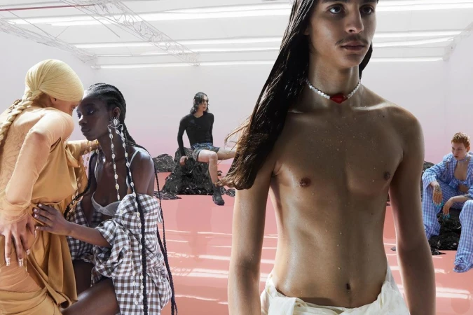 Multiple models in a bright studio with a glossy pink floor featuring a shirtless person foregrounded wearing a beaded necklace