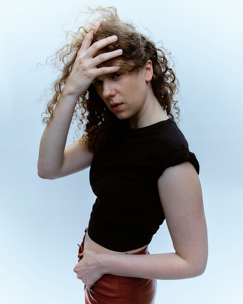 Young woman with curly hair poses in a black crop top and brown leather pants against a bright backdrop in a medium studio shot.
