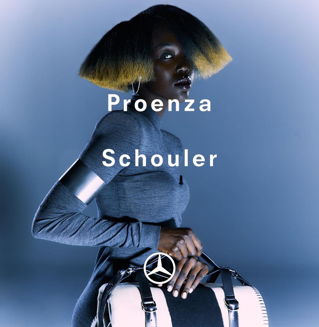 Fashion portrait of a Black woman with a dramatic hairstyle wearing a grey top and holding a duffel bag against a blue background.