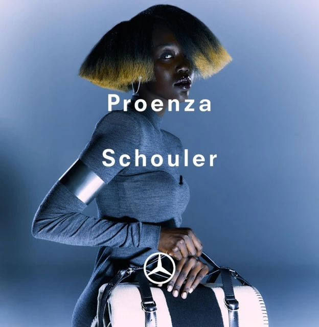 Fashion portrait of a Black woman with a dramatic hairstyle wearing a grey top and holding a duffel bag against a blue background.