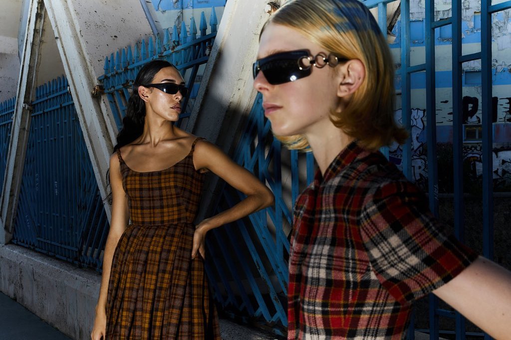 Two models wearing plaid patterned outfits and sunglasses pose outdoors against a bright blue metal gate in Paris.