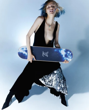 Model with blue-streaked hair holding a skateboard in a black dress against a light blue studio background