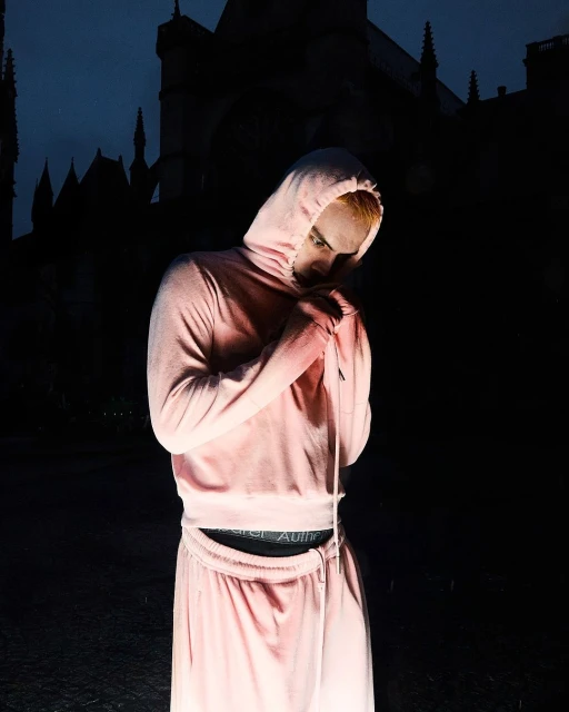 Person in a hooded sweatsuit stands outside a gothic building at night in dim light, illuminated from below in a medium shot.