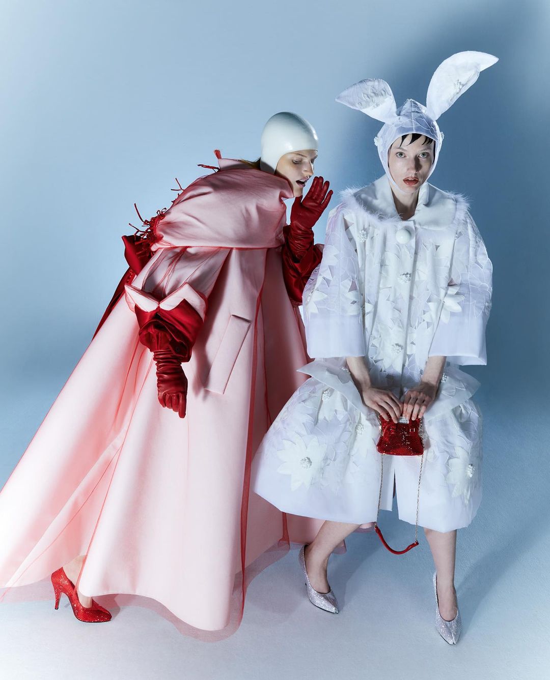 Two women in avant-garde fashion pose against a blue gradient backdrop. One wears bunny ears and a floral coat; the other wears a pink gown.