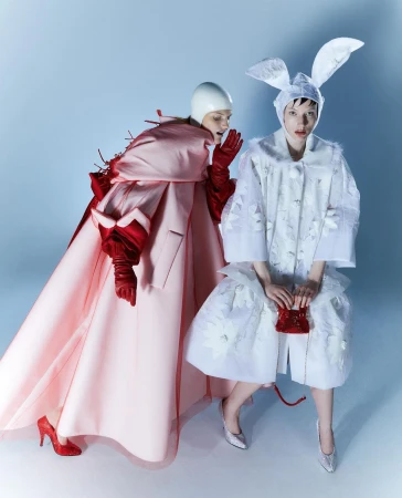 Two women in avant-garde fashion pose against a blue gradient backdrop. One wears bunny ears and a floral coat; the other wears a pink gown.