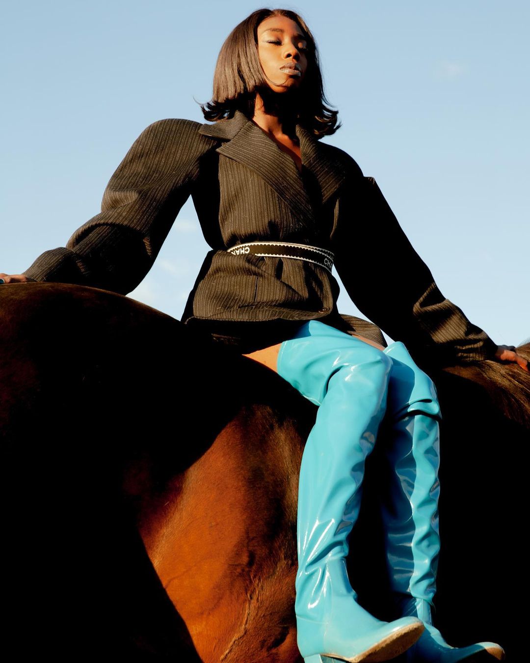 A person in a dark blazer and shiny blue boots poses on a brown horse against a clear blue sky. Fashion editorial shot outdoors.