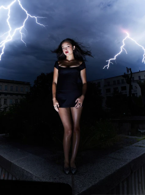 Young woman in short black dress stands on stone platform with lightning strikes behind her in a nighttime urban setting.