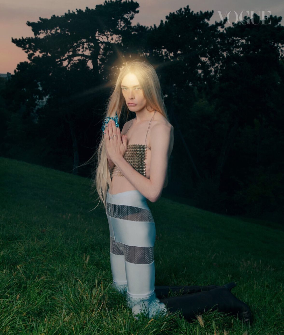 A blonde female model poses outdoors in a field wearing metallic clothing and jewelry with the Vogue logo at the top right.