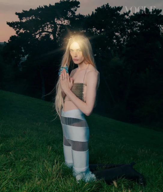 A blonde female model poses outdoors in a field wearing metallic clothing and jewelry with the Vogue logo at the top right.