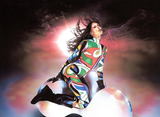 Woman in colorful patterned catsuit poses on reflective sphere against a dramatic cloudy sky with lightning strikes and bright light.