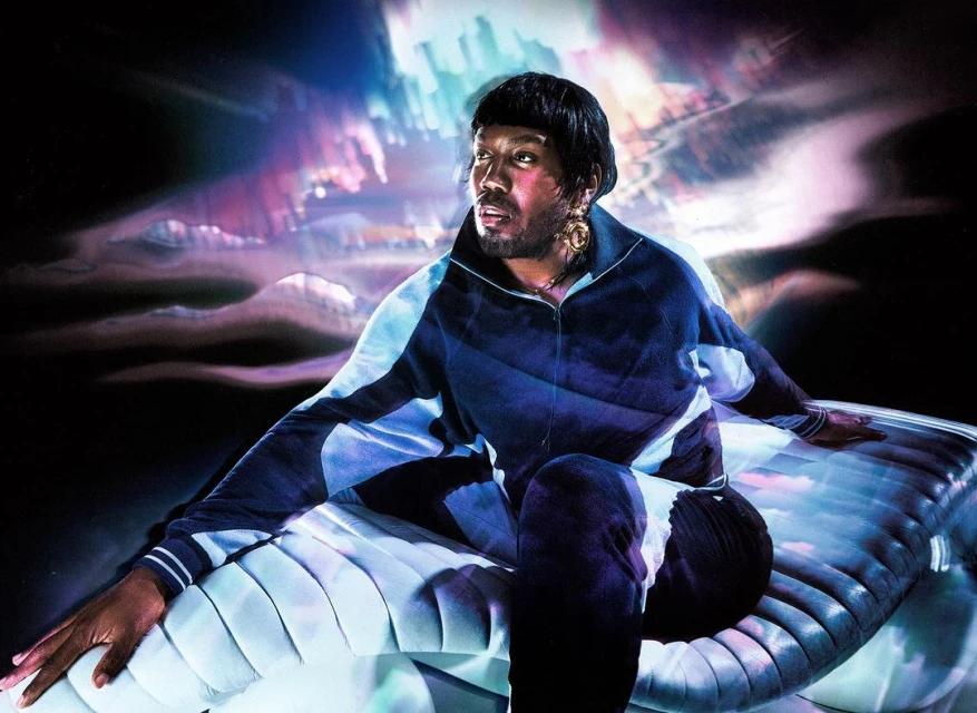 Portrait of an adult Black man in a blue tracksuit sitting on a textured surface, set against an abstract backdrop with soft lighting.