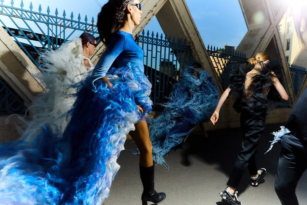 Runway fashion show on city bridge features models wearing avant-garde couture dresses in motion, with a blue sky in the background.