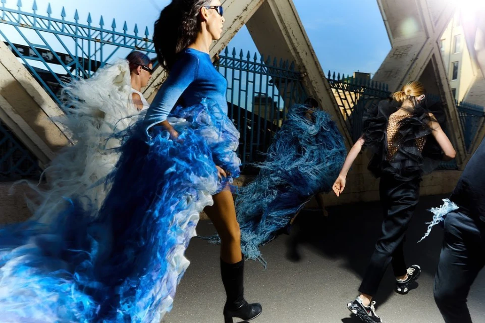 Runway fashion show on city bridge features models wearing avant-garde couture dresses in motion, with a blue sky in the background.
