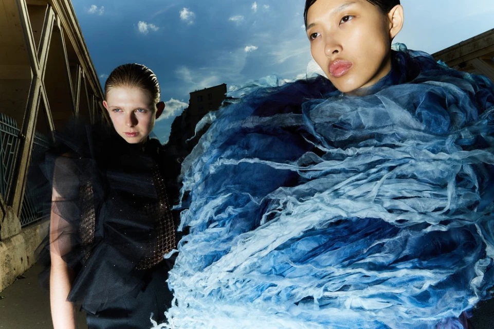 Two fashion models pose in avant-garde designs on a street with old buildings, photographed with a low angle against a cloudy sky.