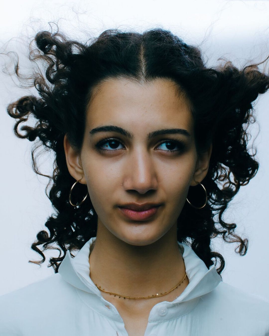 Close-up portrait of a woman with dark curly hair wearing hoop earrings and a white collared shirt against a pale background.