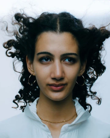 Close-up portrait of a woman with dark curly hair wearing hoop earrings and a white collared shirt against a pale background.