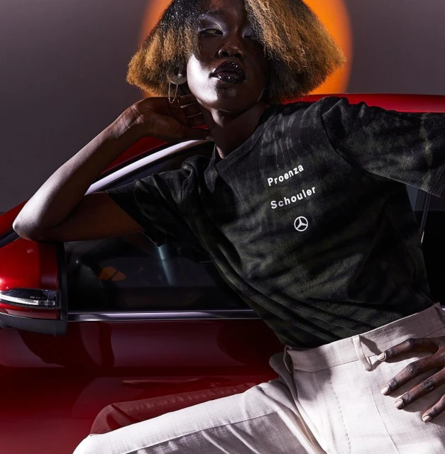 Fashion shot of a Black adult female wearing a Proenza Schouler shirt and trousers, leaning on a red car with dramatic lighting.