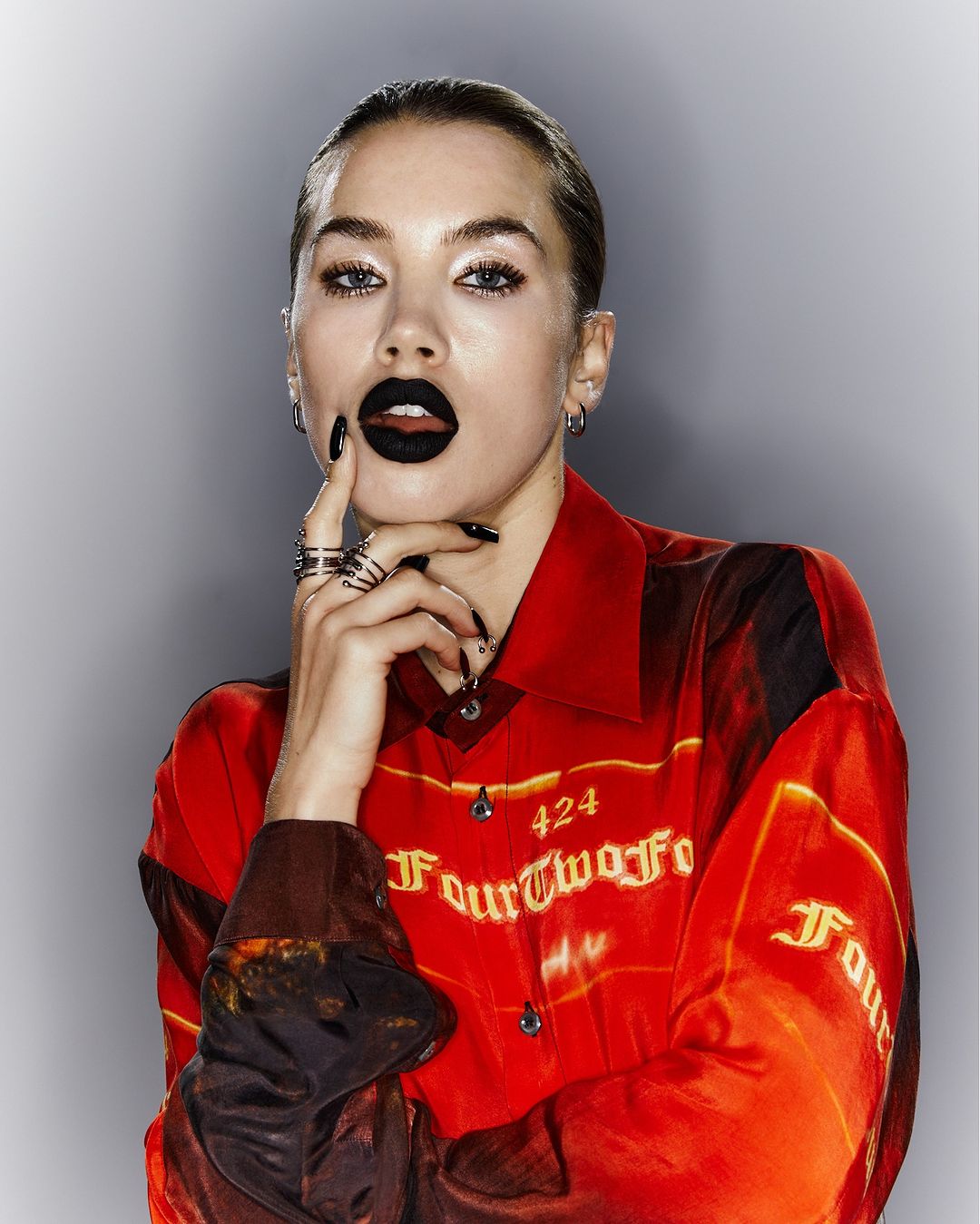 Attractive woman with black lipstick and rings poses in a red patterned shirt against a gray background in a studio shot.