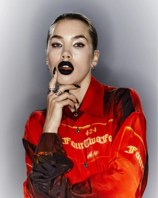 Attractive woman with black lipstick and rings poses in a red patterned shirt against a gray background in a studio shot.