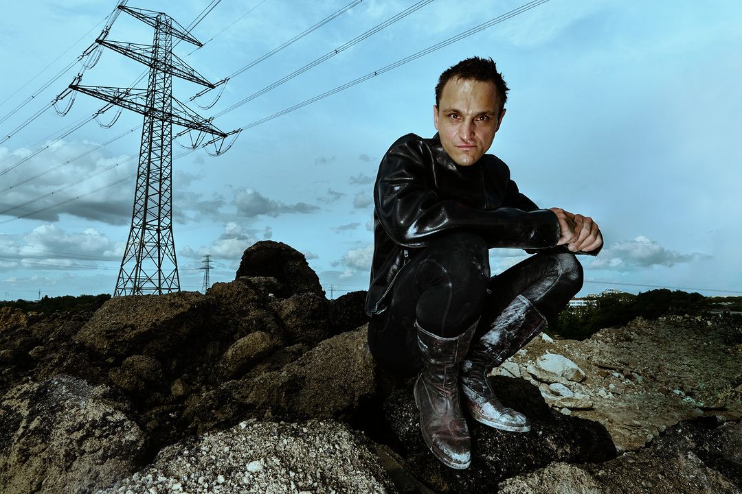An adult male in a leather jacket and boots crouches on a mound of dirt with a tall power line tower in the background under a blue sky.