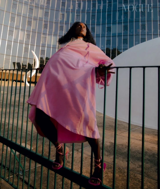 A fashionable Black woman in a pink dress and matching heels poses against a modern building on a sunny day for an outdoor fashion shot.