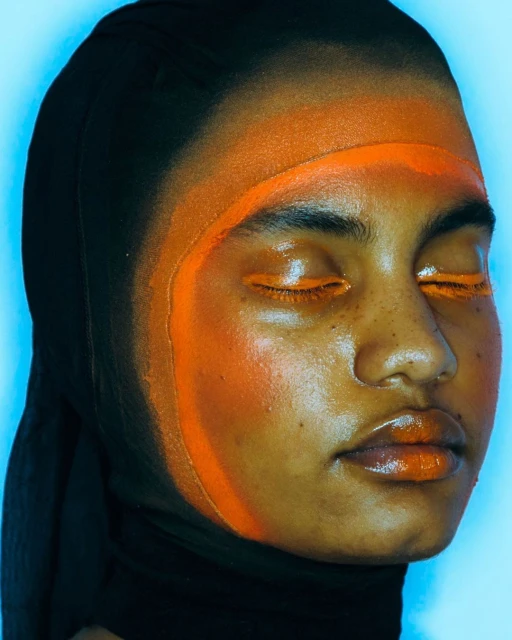 Close-up of a young person with closed eyes wearing a black head covering, accented by graphic orange paint across the face and eyelids.