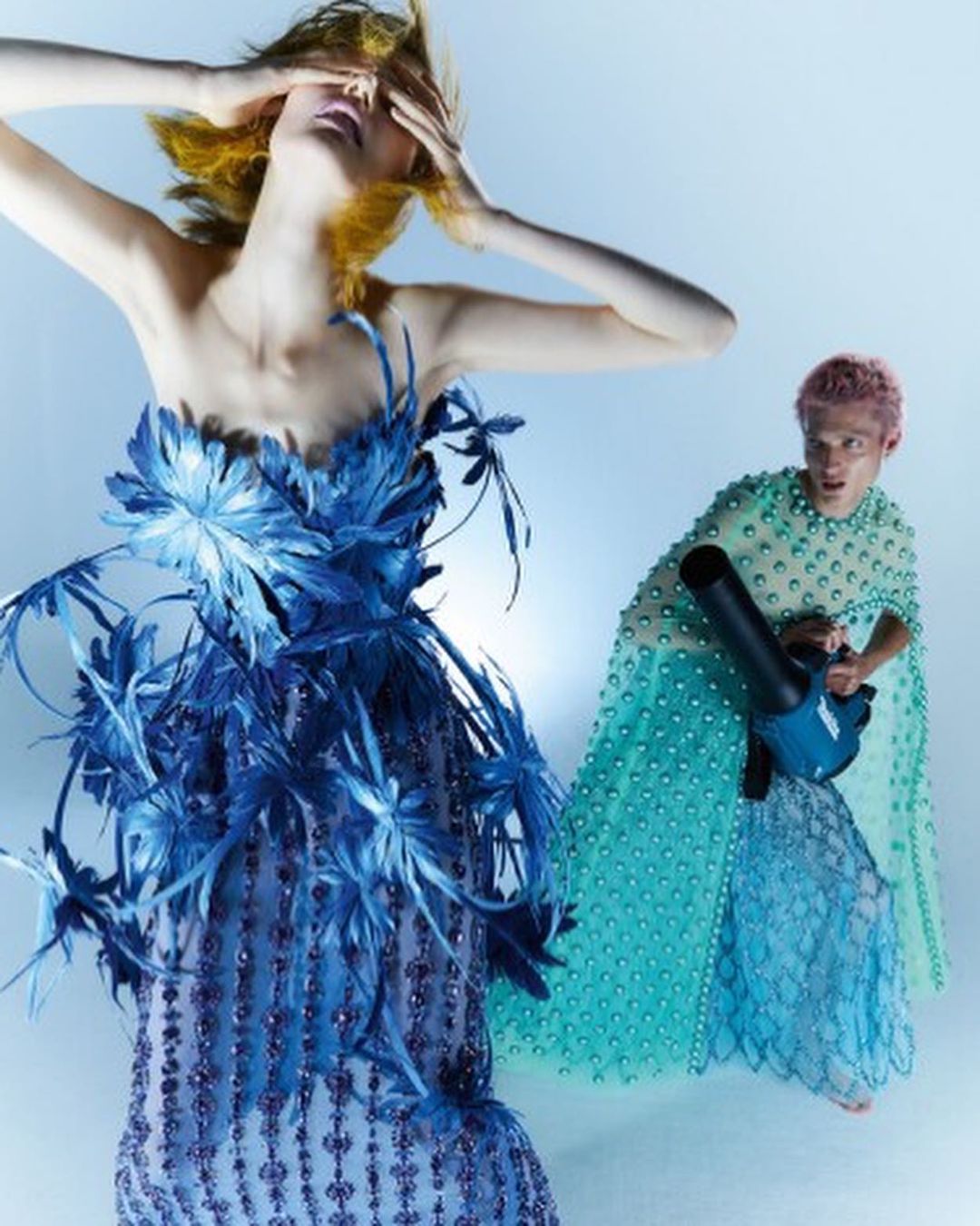 A fashion shot of a blond woman in a blue feathered dress with a man holding a leaf blower, posing in front of a light blue backdrop.