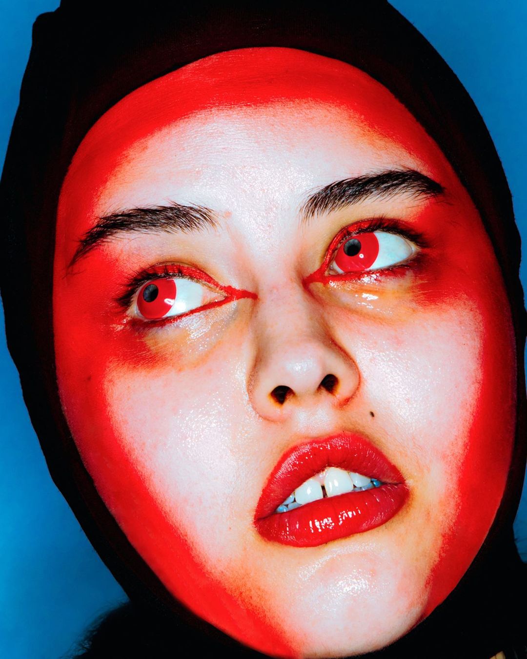 Headshot of a young woman with vibrant red face paint and matching red contacts, wearing a black head covering against a blue background.