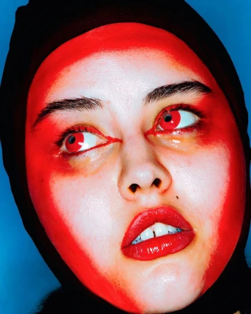 Headshot of a young woman with vibrant red face paint and matching red contacts, wearing a black head covering against a blue background.