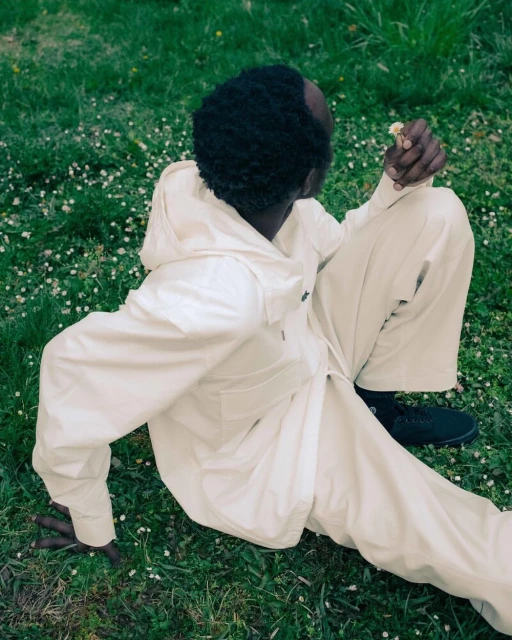 A Black adult male in light beige hooded jacket and pants sits in a grassy field with white flowers, holding a flower, looking away.