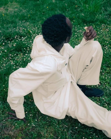 A Black adult male in light beige hooded jacket and pants sits in a grassy field with white flowers, holding a flower, looking away.