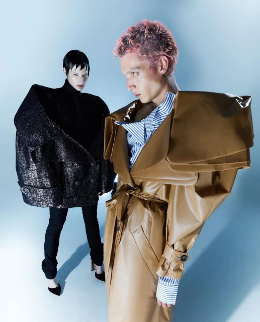 Two fashion models pose in oversized avant-garde coats in a studio setting with soft lighting and a light blue background, highlighting the garments.