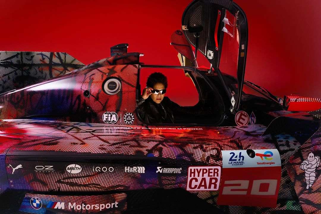 A woman wearing sunglasses is seated inside a race car, which features colorful graphics and logos, set against a vibrant red backdrop.