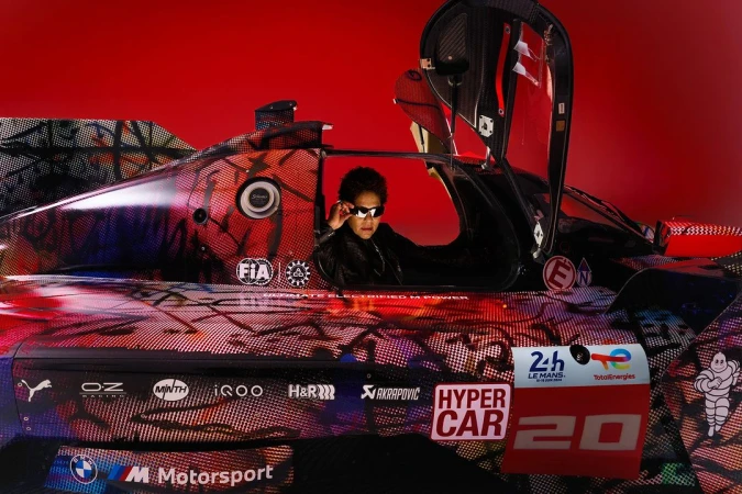 A woman wearing sunglasses is seated inside a race car, which features colorful graphics and logos, set against a vibrant red backdrop.