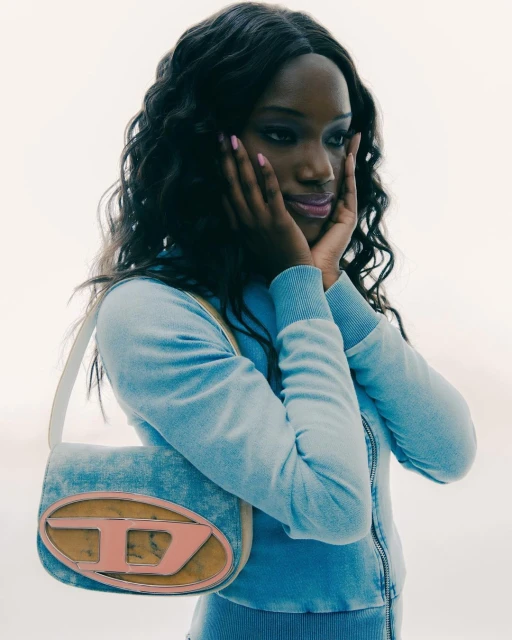 A Black woman poses with a blue denim Diesel logo bag. She has her hands on her cheeks, and a light neutral background.
