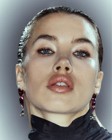 Close-up portrait of a woman with slicked-back hair and glossy lips, wearing statement earrings against a gradient grey background.
