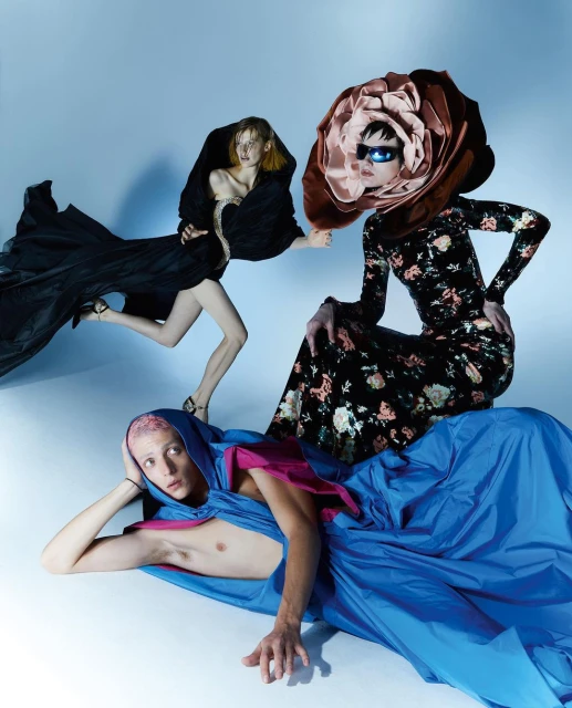 Three fashion models wearing dresses, posing in a studio with a blue backdrop. One wears a flower headpiece. High-fashion concept.