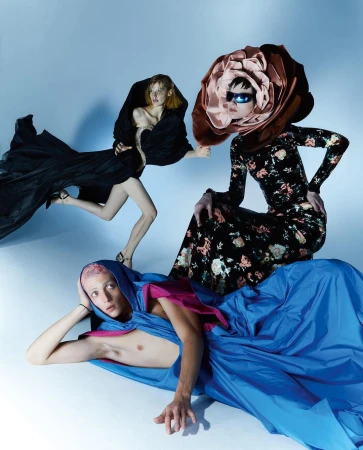 Three fashion models wearing dresses, posing in a studio with a blue backdrop. One wears a flower headpiece. High-fashion concept.