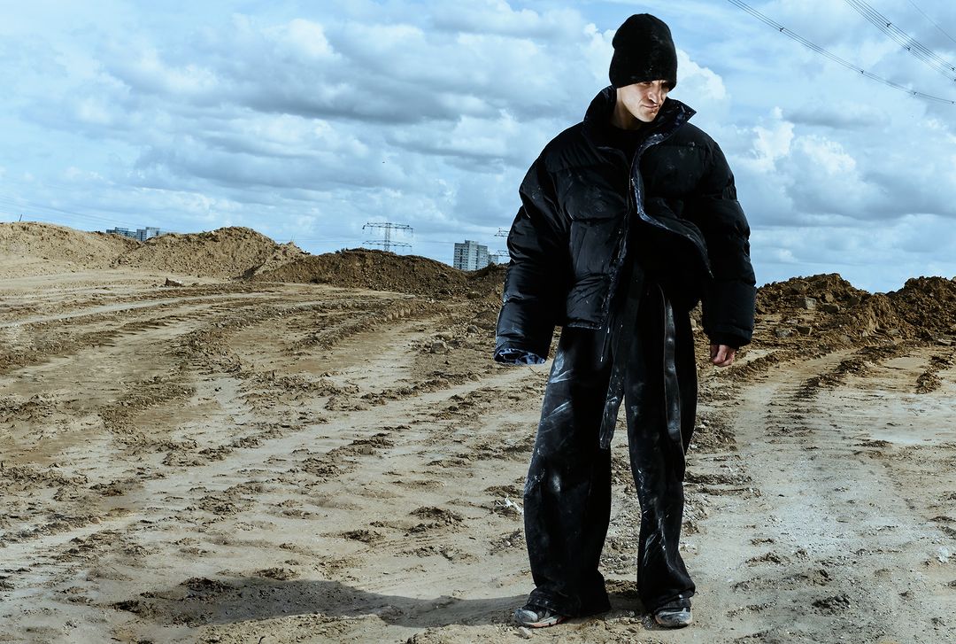 Fashion portrait of adult male in black puffy coat and beanie posing outdoors on dirt road against cloudy sky and buildings in distance.