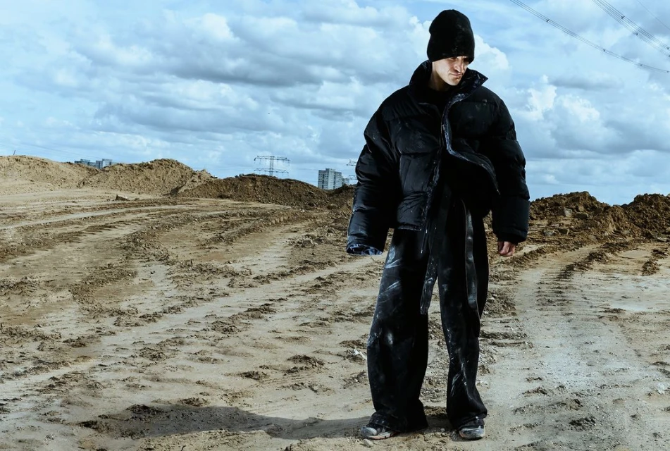 Fashion portrait of adult male in black puffy coat and beanie posing outdoors on dirt road against cloudy sky and buildings in distance.