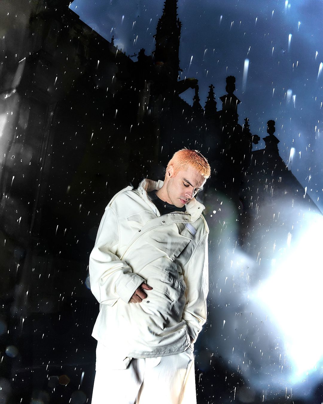 Full-length portrait of a young adult with bleached hair, standing in a white outfit against the backdrop of a gothic cathedral.