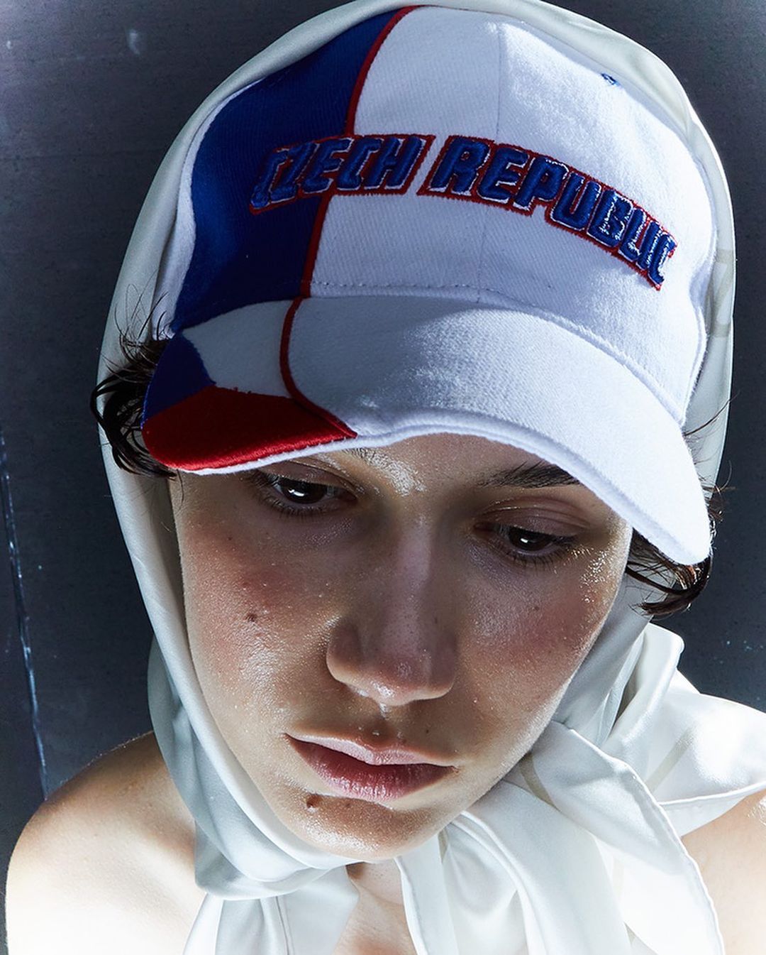 Close-up portrait of a young adult wearing a white headscarf and a baseball cap embroidered with 'Czech Republic' against a dark background.