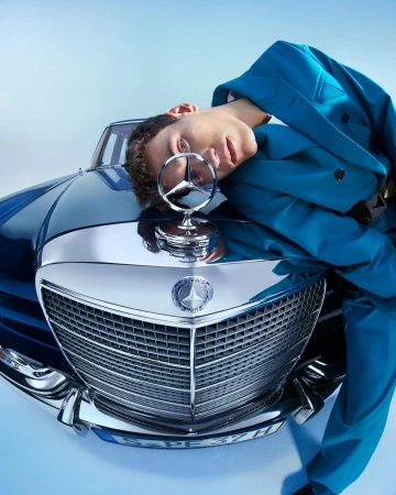 Young man in a teal suit leans on the hood of a vintage Mercedes-Benz car against a pale blue background, fashion automotive shot.