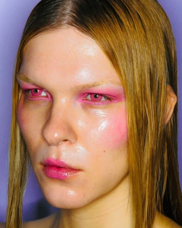 Close-up portrait of a model with glossy skin, vibrant pink eye makeup, and sleek wet-look blonde hair against purple background