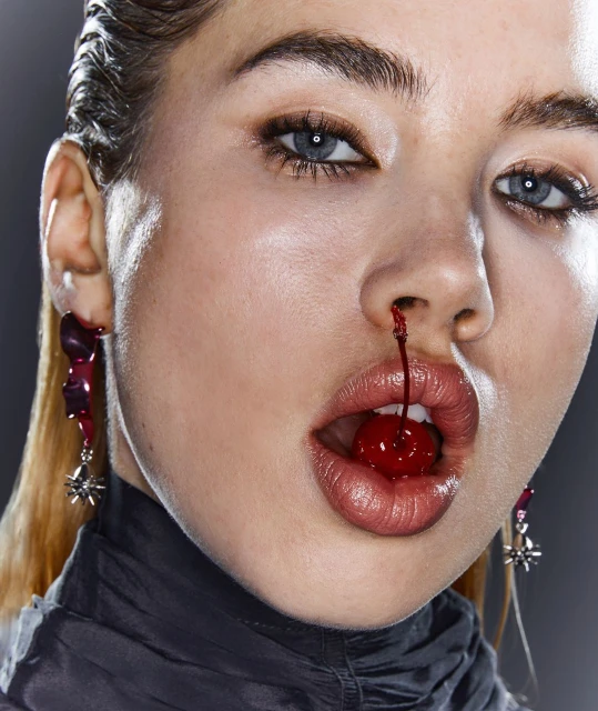 Striking beauty shot of a woman with a cherry in her mouth and juice dripping from her nose, studio lit on a gray background.