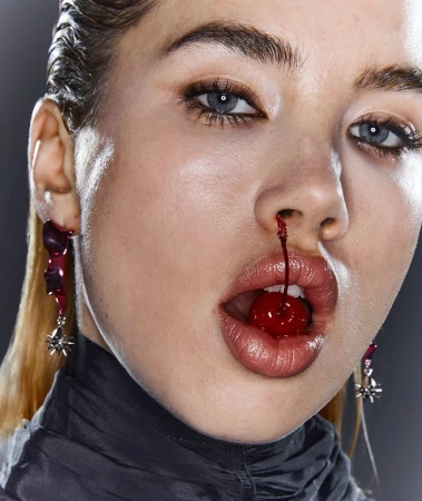 Striking beauty shot of a woman with a cherry in her mouth and juice dripping from her nose, studio lit on a gray background.