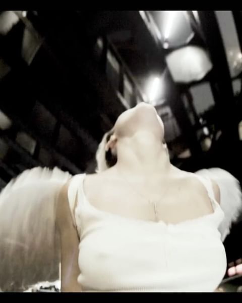 Adult female looking up wearing a white tank top and feather angel wings, shot from a low angle against a dark interior backdrop.