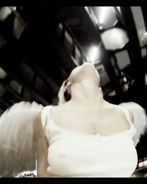Adult female looking up wearing a white tank top and feather angel wings, shot from a low angle against a dark interior backdrop.