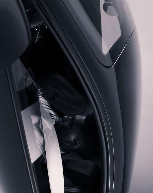 A person lies inside a sleek, dark vehicle, likely a car, viewed at an angle to show the vehicle's interior and the person's resting pose.