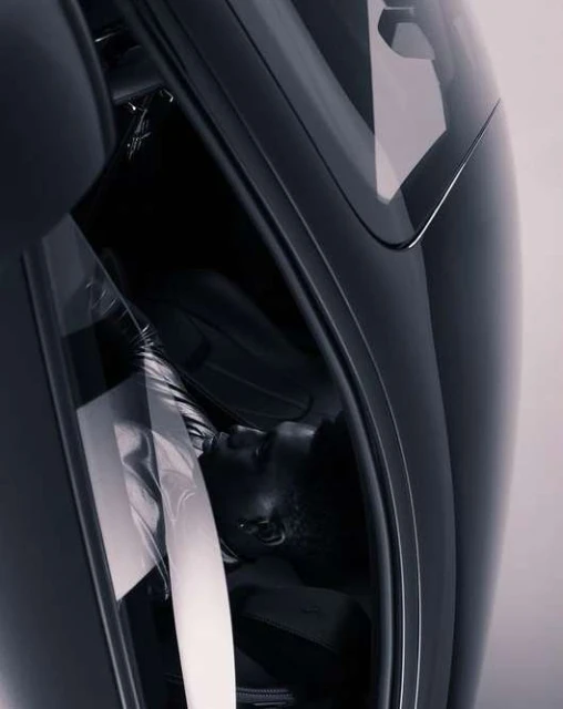 A person lies inside a sleek, dark vehicle, likely a car, viewed at an angle to show the vehicle's interior and the person's resting pose.
